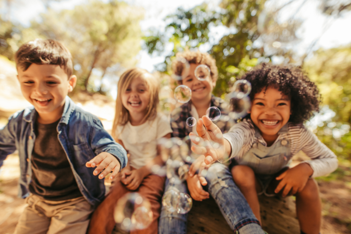 Lachende Kinder sitzen draußen in der Natur in einer Reihe und spielen mit Seifenblasen.