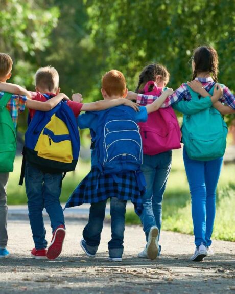 Schulkinder mit Rucksäcken auf dem Rücken laufen Arm in Arm eine Allee entlang.