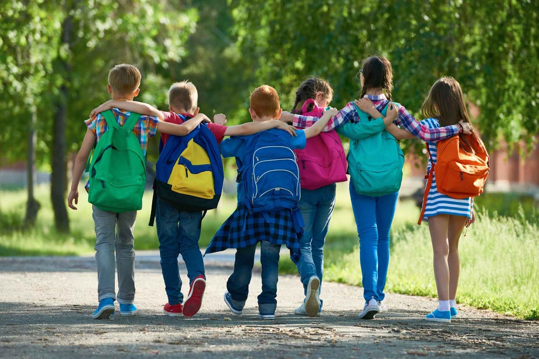 Schulkinder mit Rucksäcken auf dem Rücken laufen Arm in Arm eine Allee entlang.