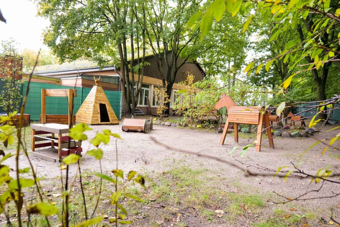 Spielplatz im Außenbereich der Kita Archenholzstraße mit Holzspielgeräten und Sandfläche, umgeben von Bäumen und Natur, ideal für Kinderaktivitäten und motorische Entwicklung.