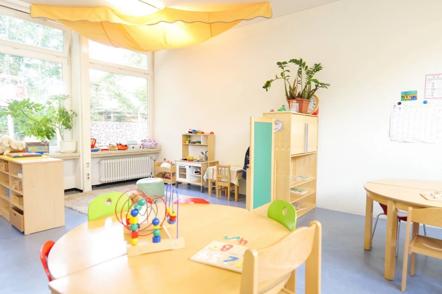 Heller, lichtdurchfluteter Gruppenraum in der Kita Redder mit Tischgruppen und einer Spielecke mit einer Kinderküche.