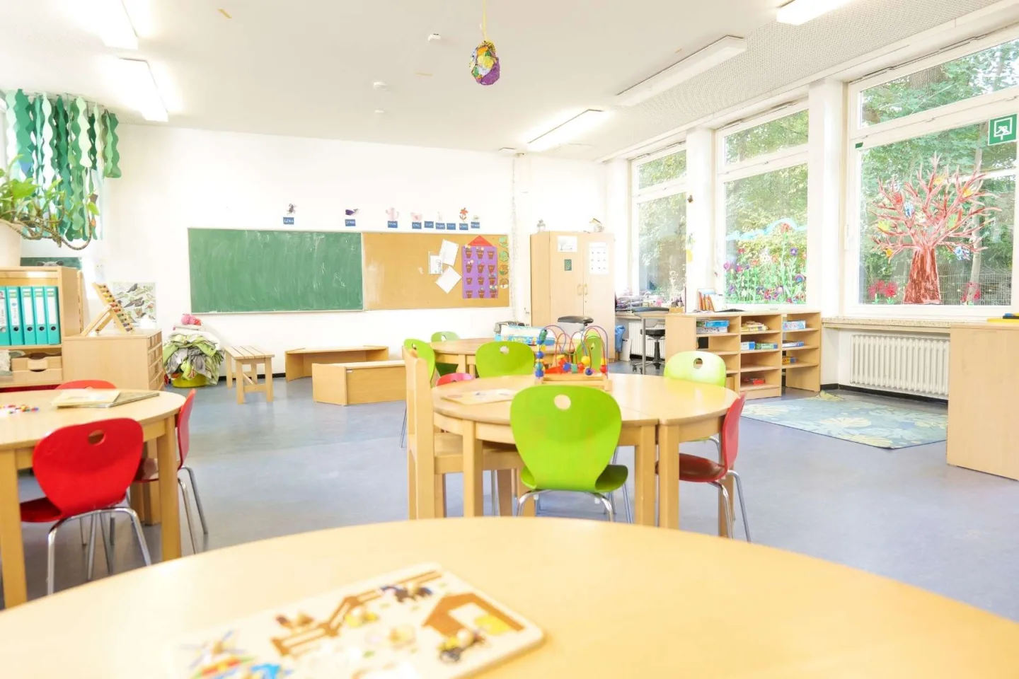 Heller, lichtdurchfluteter Gruppenraum der Kita Redder mit vielen runden Tischgruppen, Spielecken und einer großen grünen Tafel an der Wand.