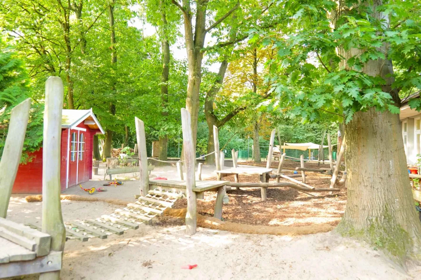 Weitläufiges, grünes Außengelände der Kita Rungwisch mit vielen Bäumen, einem Spielhaus und einem Klettergerüst aus Holz.