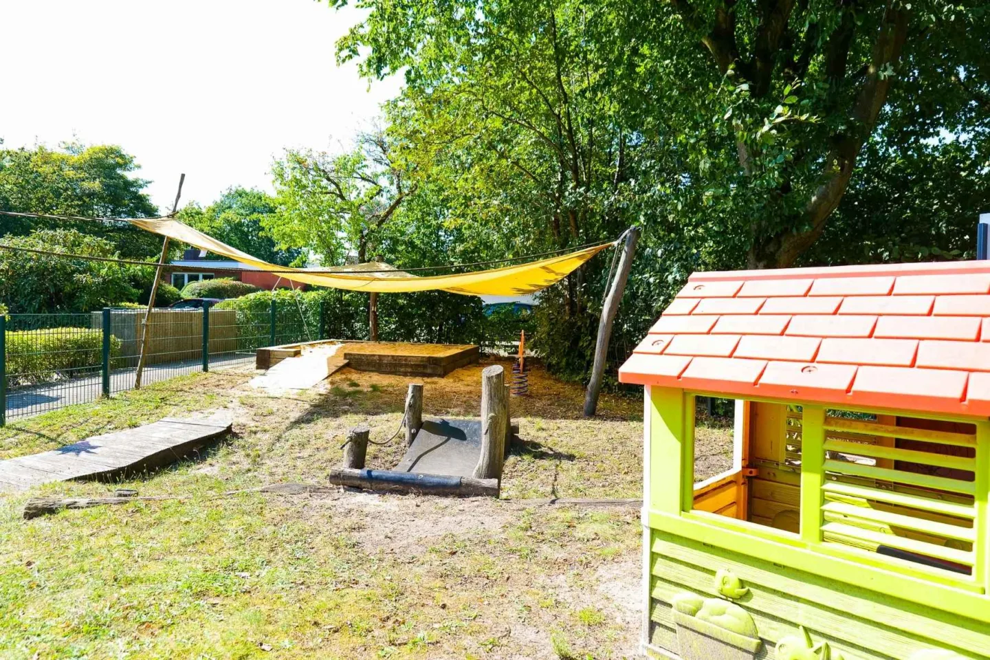 Sonniger Außenbereich in der Kita Schafgarbenweg mit einem Spielhäuschen, Klettermöglichkeiten, einem Sandkasten sowie einem gelben Sonnensegel.