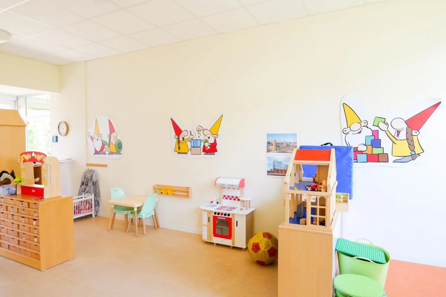 Heller, bunter Gruppenraum in der Kita Schafgarbenweg mit Spielküche, Sitzgelegenheiten, Puppenhaus und bunten Wichtelfiguren an den Wänden.
