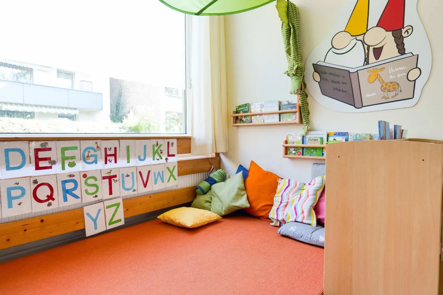 Helle, gemütliche Leseecke in der Kita Schafgarbenweg mit vielen Kissen und Decken. An der Wand hängen Bücherregale sowie ein großes Wandbild von zwei Wichteln die Lesen.