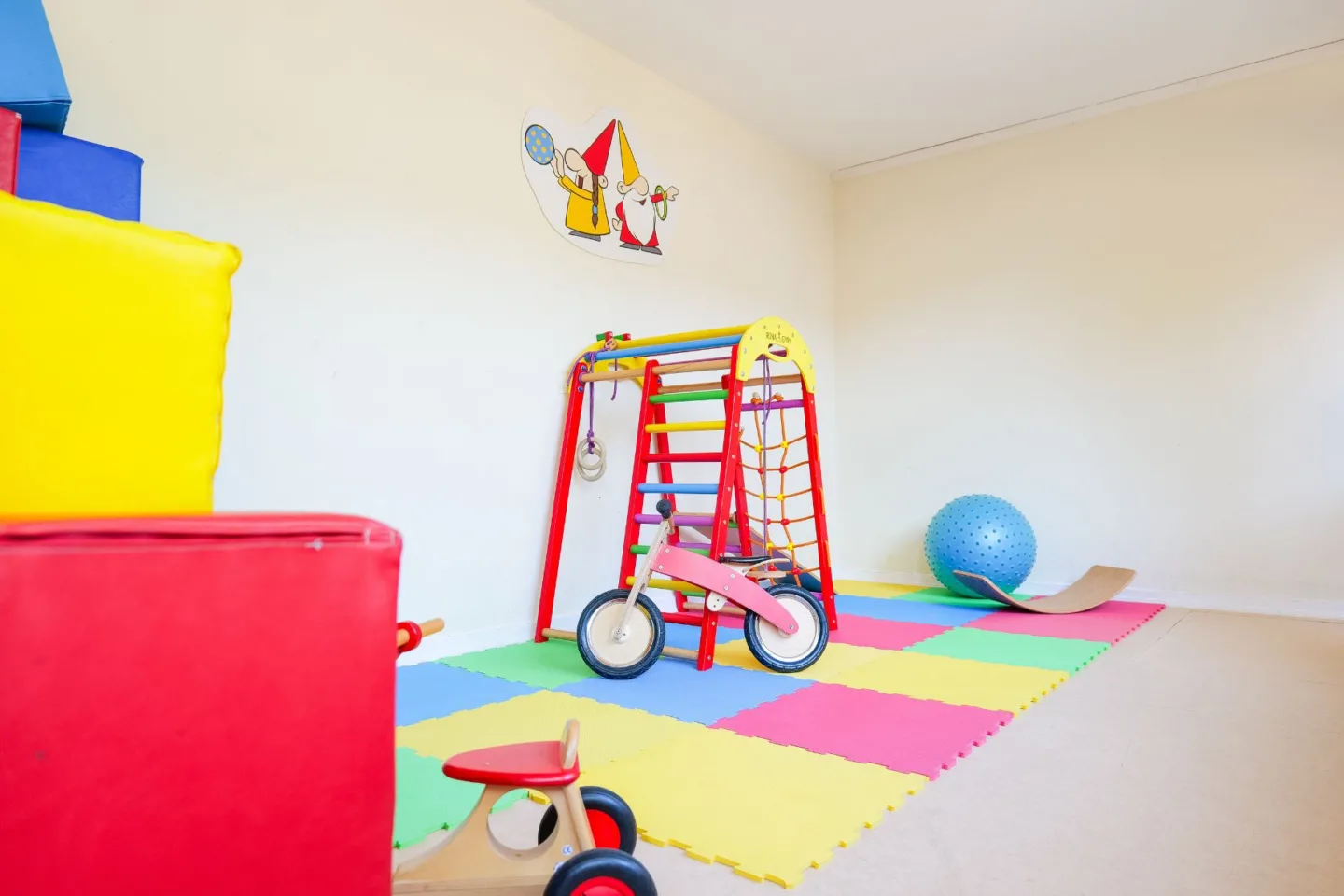 Heller, bunter Gruppenraum in der Kita Schafgarbenweg mit einem Indoor-Klettergerüst, Indoor-Kinderfahrzeugen, Gymnastikbällen und einem Gleichgewichtsboard.