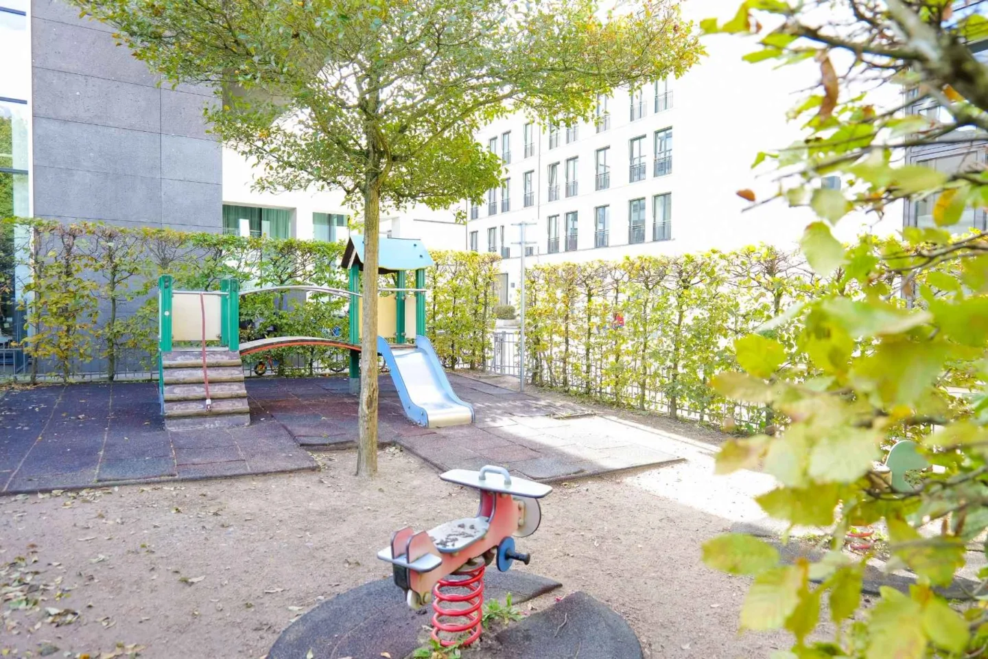 Grüner, von Bäumen umgebener Spielplatz der Kita Völckersstraße mit einer Wippfigur und einem bunte Klettergerüst mit Rutsche.