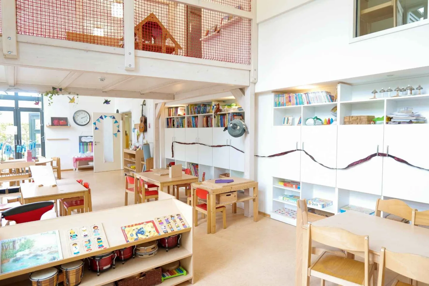 Heller, lichtdurchfluteter Gruppenraum in der Kita Völckersstraße mit Tischgruppen, Regalen mit vielen Büchern, Musikinstrumenten und bunten Basteleien an der Wand.